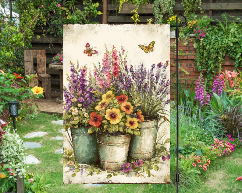 May include: A garden flag with a watercolour painting of colourful flowers in metal pots. The flowers include purple, pink, yellow, and orange blooms. Two butterflies are depicted above the flowers. The flag is displayed in a garden setting.
