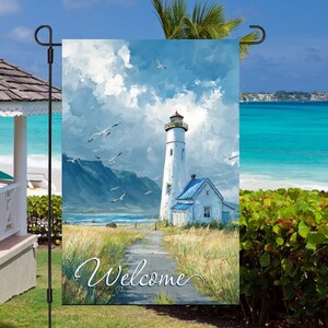 May include: A garden flag featuring a coastal scene with a white lighthouse, blue roof, and the word "Welcome" in script. The artwork depicts a path leading to the lighthouse, with seagulls flying in a blue sky. The flag is displayed outdoors.