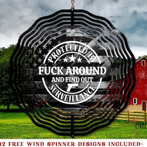 May include: Black metal wind spinner with a circular design. The center features a distressed American flag with the text "PROTECTED BY FUCK AROUND AND FIND OUT SURVEILLANCE." The background shows a red barn and green field.
