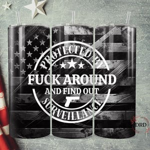 May include: Three stainless steel tumblers with a black and white American flag design. The tumblers feature the text "PROTECTED BY FUCK AROUND AND FIND OUT SURVEILLANCE" in a white circular design. Each tumbler has a clear straw.