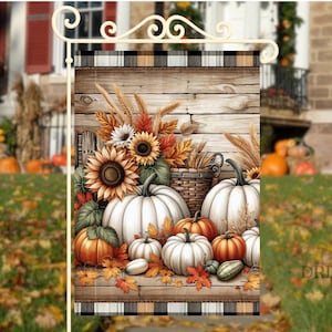 Puede incluir: Una bandera de jardín decorativa con una escena de cosecha de otoño. El diseño incluye calabazas blancas y naranjas, girasoles, trigo y hojas de otoño. El fondo tiene un diseño de tablones de madera con un borde a cuadros blanco y negro.