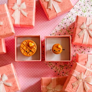May include: A collection of square, textured coral-colored gift boxes with light pink ribbon bows. Two open boxes reveal miniature food-themed jewelry: one features a round waffle with a heart-shaped butter pat, and the other a circular pancake.