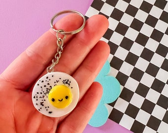 Puede incluir: Un llavero de huevo frito blanco y amarillo con una cara sonriente. El huevo está rodeado de purpurina negra. El llavero está unido a una cadena plateada.