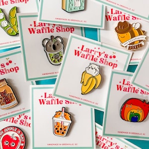May include: Assortment of enamel pins featuring various cartoon characters and designs. The pins are all handmade in Greenville, SC and feature a variety of colors and themes. The pins are displayed on cards with the text "Larry's Waffle Shop" printed on them.