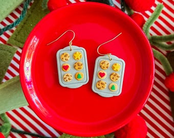 May include: A pair of dangle earrings shaped like rectangular gray baking sheets, each featuring six small cookies with brown textures, a yellow star, a red heart, and a green triangle, suspended from gold-toned metal earring hooks.