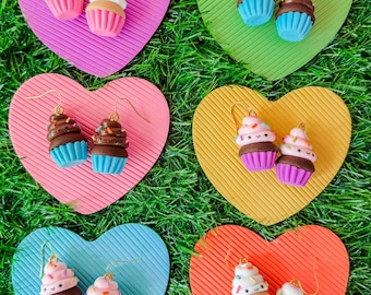 Puede incluir: Seis pares de pendientes de cupcake, cada uno con un diseño único. Los pendientes se exhiben sobre bases en forma de corazón en varios colores, incluyendo rosa, verde, amarillo, azul y naranja. Los cupcakes tienen diferentes glaseados y chispas.