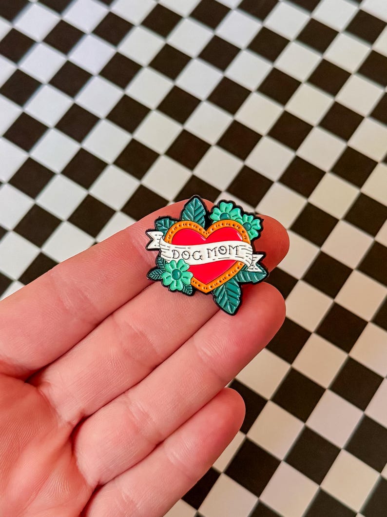 May include: Red heart with a banner that reads "Dog Mom" surrounded by green leaves and flowers. The pin is made of enamel and has a black outline.