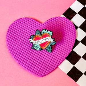 May include: A pink heart-shaped pin cushion with a red heart enamel pin featuring the text "Dog Mom" and floral accents.