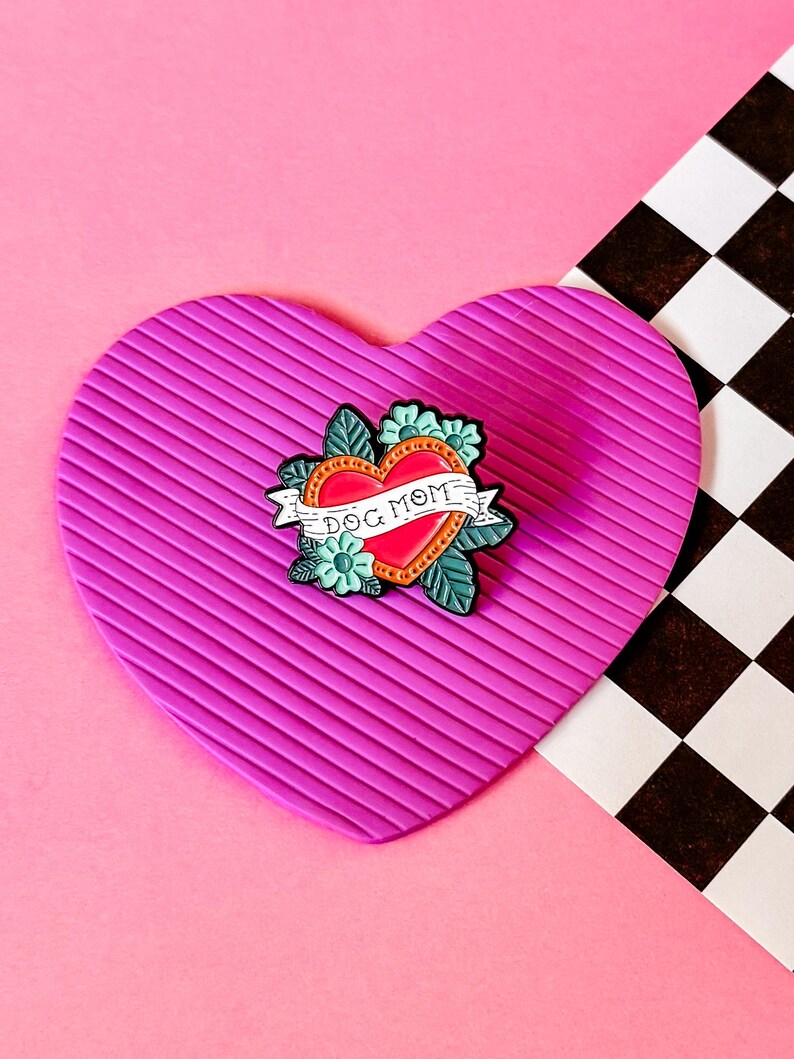May include: A pink heart-shaped pin cushion with a red heart enamel pin featuring the text "Dog Mom" and floral accents.