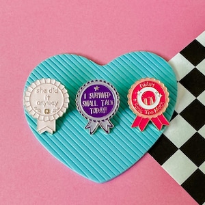 May include: Three enamel pins with white, purple, and pink backgrounds. The white pin says "she did it anyway". The purple pin says "I survived small talk today!" The pink pin says "Didn't panic too much".