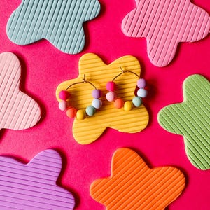 May include: Gold hoop earrings with colorful beaded accents, displayed on a yellow flower-shaped base. The earrings feature pink, purple, blue, and yellow beads. The background is a vibrant pink, with flower-shaped decorations in various colors.