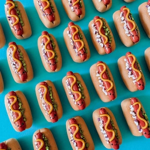 Op de afbeelding: Een verzameling van 20 miniatuur hotdogs gemaakt van polymer clay. Elke hotdog is versierd met gele mosterd, rode relish en witte uien. De hotdogs hebben een schattig, cartoonachtig gezicht met ogen en een glimlach.