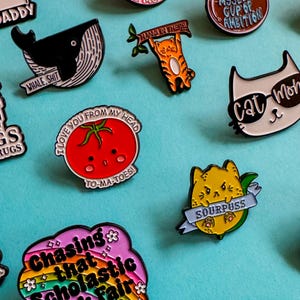 May include: A collection of enamel pins featuring various designs, including a whale, a tomato, a cat, a frog, a lemon, and a rainbow with the text "Chasing that Scholastic Book Fair high".
