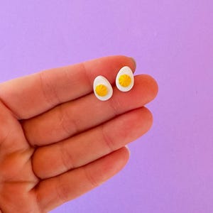 May include: A pair of white and yellow egg-shaped stud earrings. The earrings feature a white egg white with a bright yellow yolk in the center. The earrings are small and delicate, perfect for everyday wear.