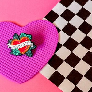 May include: A pink heart-shaped background with a black and white checkered pattern in the background. A green, red, and black enamel pin with a heart and the words "Dog Mom" is on the pink heart.