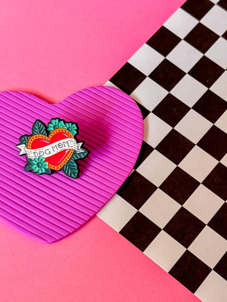 May include: A pink heart-shaped background with a black and white checkered pattern in the background. A green, red, and black enamel pin with a heart and the words "Dog Mom" is on the pink heart.