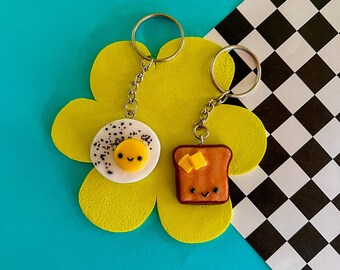 Puede incluir: Un par de llaveros con un huevo frito y una rebanada de tostada. El huevo tiene una carita linda con una yema amarilla y motas negras. La tostada tiene una corteza marrón y una carita feliz con una porción de mantequilla amarilla. Los llaveros están unidos a cadenas y anillos plateados.