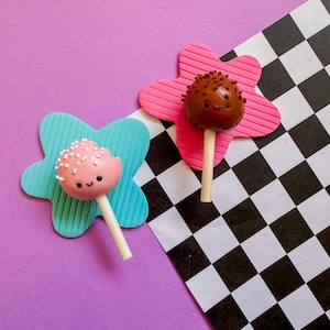 May include: Two cake pops on sticks, one pink with white sprinkles and the other chocolate with brown sprinkles. They rest on flower-shaped platforms, one blue and one pink, against a purple background and a black and white checkered pattern.