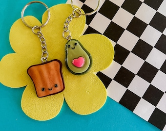 Puede incluir: Un par de llaveros con un aguacate y una rebanada de tostada de dibujos animados. El aguacate es verde con un corazón rosa y una cara sonriente. La tostada es marrón con una cara sonriente. Los llaveros están colgados de un fondo amarillo en forma de flor.
