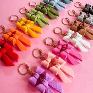 May include: A collection of colorful bow-shaped keychains with gold key rings. The bows are made of a textured material and come in a variety of colors, including red, orange, yellow, green, blue, purple, pink, and white.