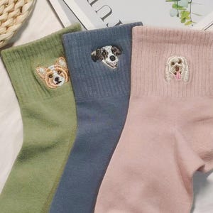 May include: Three pairs of ribbed ankle socks in sage green, slate blue, and soft pink. Each sock features a small, detailed embroidered dog face on the upper cuff, showcasing different breeds with distinct facial markings and colours.