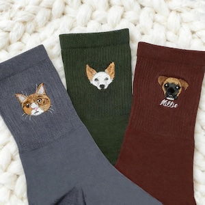 Könnte beinhalten: Drei Paar Socken mit gestickten Tiergesichtern. Ein Paar ist dunkelgrau mit einem Katzengesicht, ein anderes ist olivgrün mit einem Hundegesicht, und das dritte ist weinrot mit einem Hundegesicht und dem Namen "Millie".