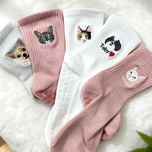 Puede incluir: Un conjunto de cuatro calcetines de canalé en gris claro, rosa empolvado y blanco. Cada calcetín presenta una cara de animal bordada, incluyendo un perro y tres gatos. Un calcetín blanco muestra el nombre Toby con un corazón rojo. Las suelas tienen agarres.