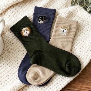 May include: Three pairs of crew socks with embroidered dog faces. One pair is dark green, one is navy blue, and one is tan. The dog breeds are a golden retriever, a black and tan dog, and a Yorkie. The socks are on a white knitted blanket.