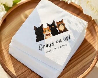 May include: A stack of white paper napkins with a cartoon illustration of four cats. The cats are black, orange, and brown, with the text "Drinks on us!" and names below. The napkins are on a wooden tray.