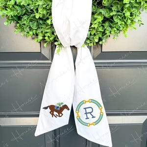 以下が含まれることがあります： 白の生地のスカーフ。競馬のデザインと、青と金の円の中に文字「R」があります。スカーフは緑のリースに掛けられています。馬は茶色で、緑と青のジョッキーがいます。