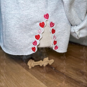 May include: A light gray sweatshirt with a decorative embroidered design of red and pink hearts. The hearts are arranged in a vertical pattern along the bottom edge of the sweatshirt. The design is on a light beige fabric.