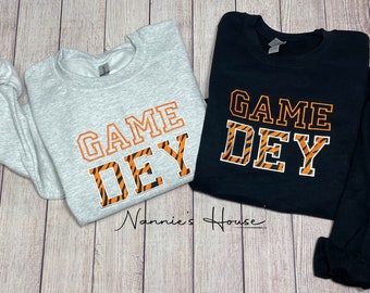 Può includere: Due felpe con la scritta "GAME DAY" stampata su di esse. Il testo è in un motivo a strisce di tigre nero e arancione. Una felpa è grigia e l'altra è nera.