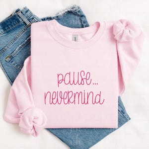 May include: A light pink long-sleeve crewneck sweatshirt featuring the dark pink embroidered text "pause... nevermind" in a cursive font, layered over a pair of blue denim jeans with a visible pocket and seam detail.