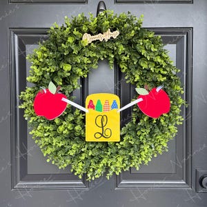 May include: A green wreath with a wooden sign that says "Nannie's House". The wreath is decorated with two red apple cutouts and a yellow rectangle with a crayon box design and the letter "L".