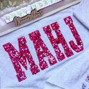 Op de afbeelding: Een lichtgrijs sweatshirt met het woord "MAHJ" in grote, roze letters met pailletten. Een houten bord met de tekst "Nannie's House" staat boven het sweatshirt, samen met een set Mahjong-tegels. Het sweatshirt is opgevouwen.