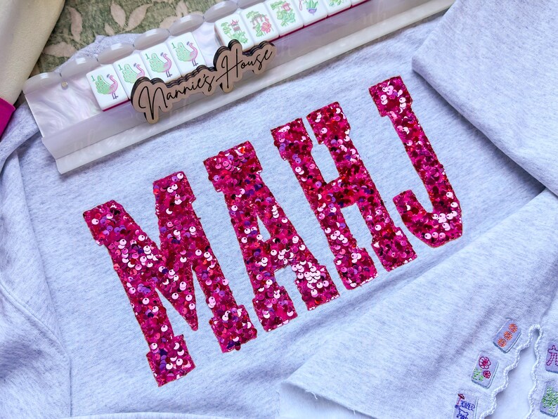 Op de afbeelding: Een lichtgrijs sweatshirt met het woord "MAHJ" in grote, roze letters met pailletten. Een houten bord met de tekst "Nannie's House" staat boven het sweatshirt, samen met een set Mahjong-tegels. Het sweatshirt is opgevouwen.