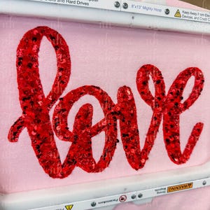 May include: A light pink fabric with the word "love" embroidered in red sequins. The embroidery is in a cursive font. The fabric is held in an embroidery hoop. The hoop is 8"x13".