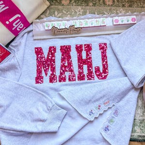 Op de afbeelding: Een lichtgrijs sweatshirt met het woord "MAHJ" in grote, sprankelende roze letters. De mouwen hebben decoratieve borduursels. Een houten dienblad met Mahjong-tegels en een draagtas zijn ook zichtbaar.