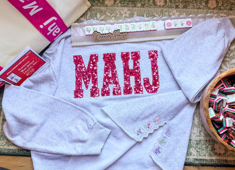 Op de afbeelding: Een lichtgrijs sweatshirt met het woord "MAHJ" in grote, sprankelende roze letters. De mouwen hebben decoratieve borduursels. Een houten dienblad met Mahjong-tegels en een draagtas zijn ook zichtbaar.