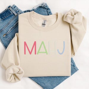 May include: A cream-colored sweatshirt with the word "MAHJ" embroidered in pink, green, light gray, and blue. The sweatshirt is folded on top of a pair of blue jeans.