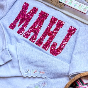 Op de afbeelding: Een lichtgrijs sweatshirt met het woord "MAHJ" in grote, felroze pailletten. De sweatshirt heeft geborduurde patches op de mouwen en wordt gecombineerd met Mahjong-tegels en een houten bord met "Nannie's House".
