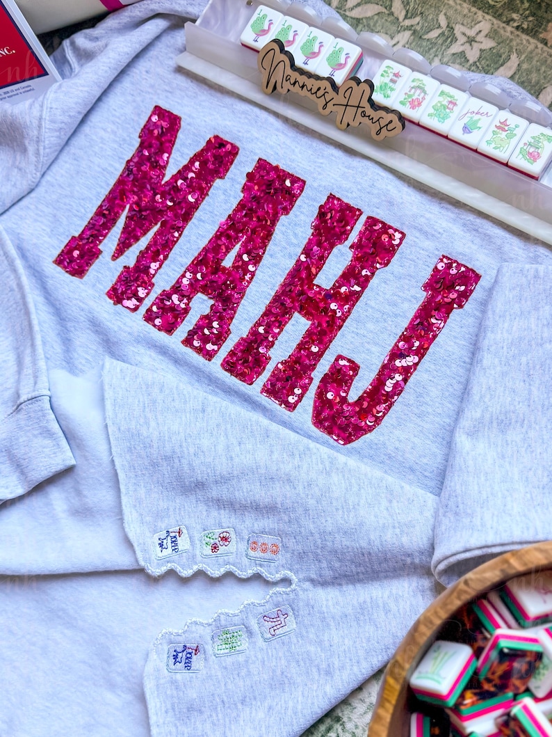 Op de afbeelding: Een lichtgrijs sweatshirt met het woord "MAHJ" in grote, felroze pailletten. De sweatshirt heeft geborduurde patches op de mouwen en wordt gecombineerd met Mahjong-tegels en een houten bord met "Nannie's House".