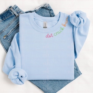 May include: A light blue crew neck sweatshirt with the text "dot crack bar" in rainbow colors. The sweatshirt is folded over a pair of blue jeans.