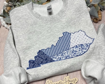 Könnte beinhalten: Ein graues Sweatshirt mit einer blauen gestickten Umrandung des Bundesstaates Kentucky. Die Umrandung besteht aus verschiedenen blauen gemusterten Stoffen, darunter Gingham, Blumen und Paisley.