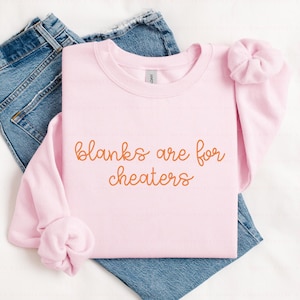 May include: A light pink sweatshirt with the text "blanks are for cheaters" in a cursive font. The sweatshirt is folded on top of a pair of blue jeans. A matching pink scrunchie is on the sleeve.
