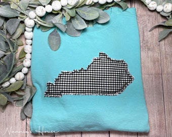 Könnte beinhalten: Ein türkisfarbenes T-Shirt mit einem schwarzen und weißen Gingham-Umriss des Bundesstaates Kentucky. Der Umriss ist mit einem ausgefransten Rand angenäht.