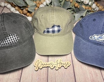 Op de afbeelding: Drie baseballpetten met een patch in de vorm van de omtrek van Kentucky. De patches zijn gemaakt van stof en hebben verschillende patronen. De petten zijn in verschillende kleuren: zwart, kaki en blauw.