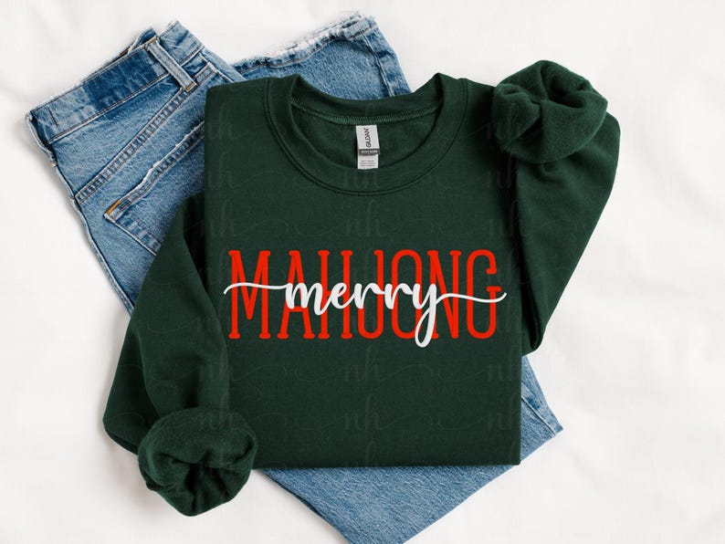 May include: A dark green sweatshirt with the words "MAHJONG merry" in red and white lettering. The sweatshirt is folded on top of a pair of blue jeans. The sleeves are rolled up.