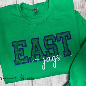 May include: Green sweatshirt with the word "EAST" in large, navy blue and green letters with a sparkly finish. Below the word "EAST" is the word "jags" in white cursive writing.