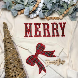 Pode incluir: Uma camisola branca de manga comprida com a palavra "MERRY" bordada em xadrez vermelho e verde. Um laço vermelho e verde xadrez está bordado abaixo da palavra. A camisola está sobre uma superfície de madeira com uma pequena árvore decorativa e uma placa de madeira que diz "Nannies House".